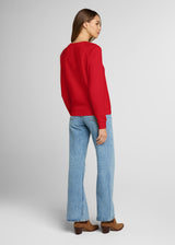 Woman wearing a red sweater and blue jeans on a light gray background. Back.