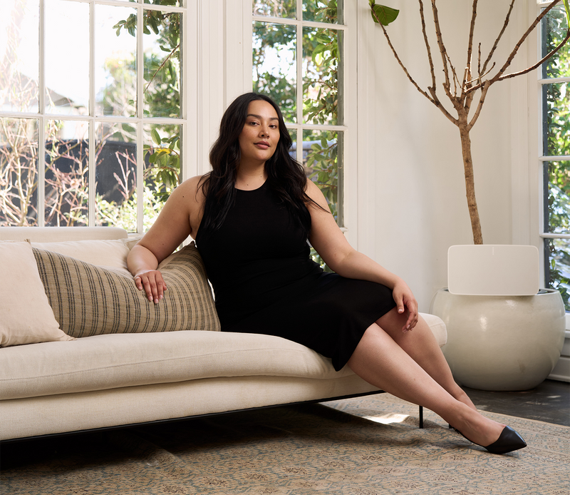 Woman in black racerback midi dress sitting on a couch in an elegant living room during the day wearing black heals. 