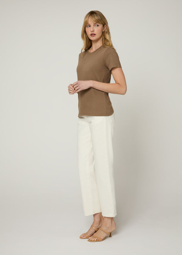 Side profile of woman in untucked brown t-shirt wearing white denim and nude heels. Her arms are together at her waist. 