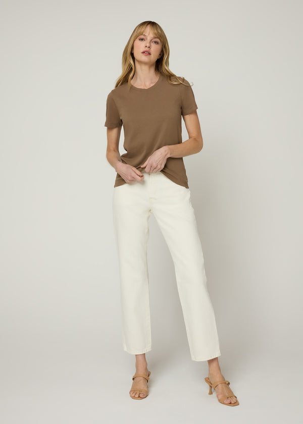Woman in brown t-shirt and white denim with nude heals. Her hands are gently tugging on the bottom of her untucked t-shirt.