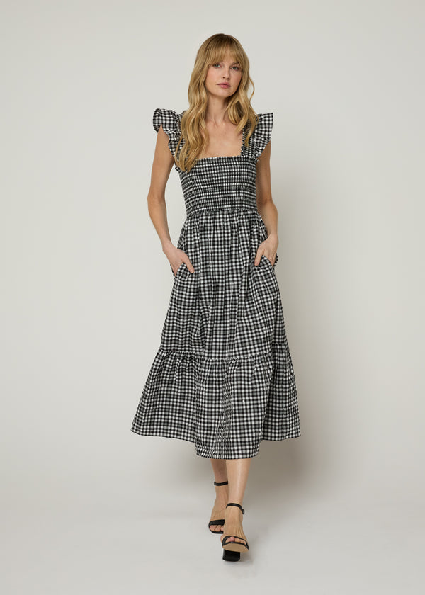 Woman in black checkered print midi dress with smocked bodice and smocked flounce shoulders with a flowy tiered skirt wearing black heals and putting her hands in dress side pockets. Detail.