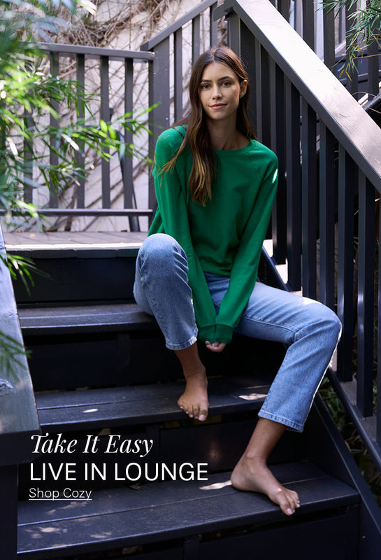 Woman in green sweatshirt and blue jeans sitting on an outdoor patio. 