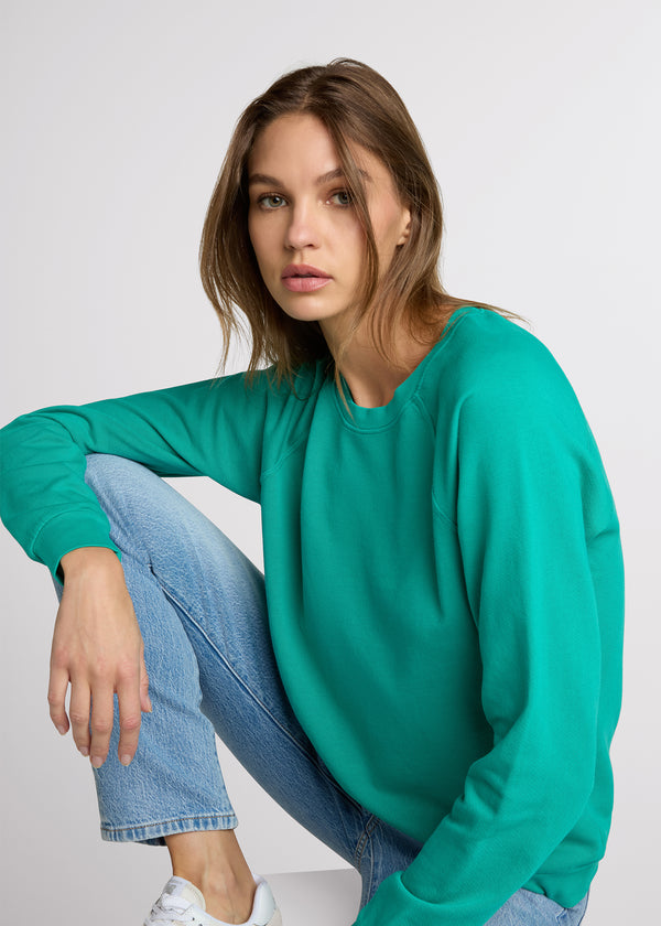 Woman in green sweatshirt and light denim sitting on a white wooden block with one hand on her knee and the other at her side. 