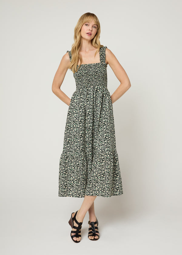 Woman in black floral midi flowy dress with a smocked bodice and smocked ruffle tank. Her hands behind her back and legs crossed in black  wedges. 