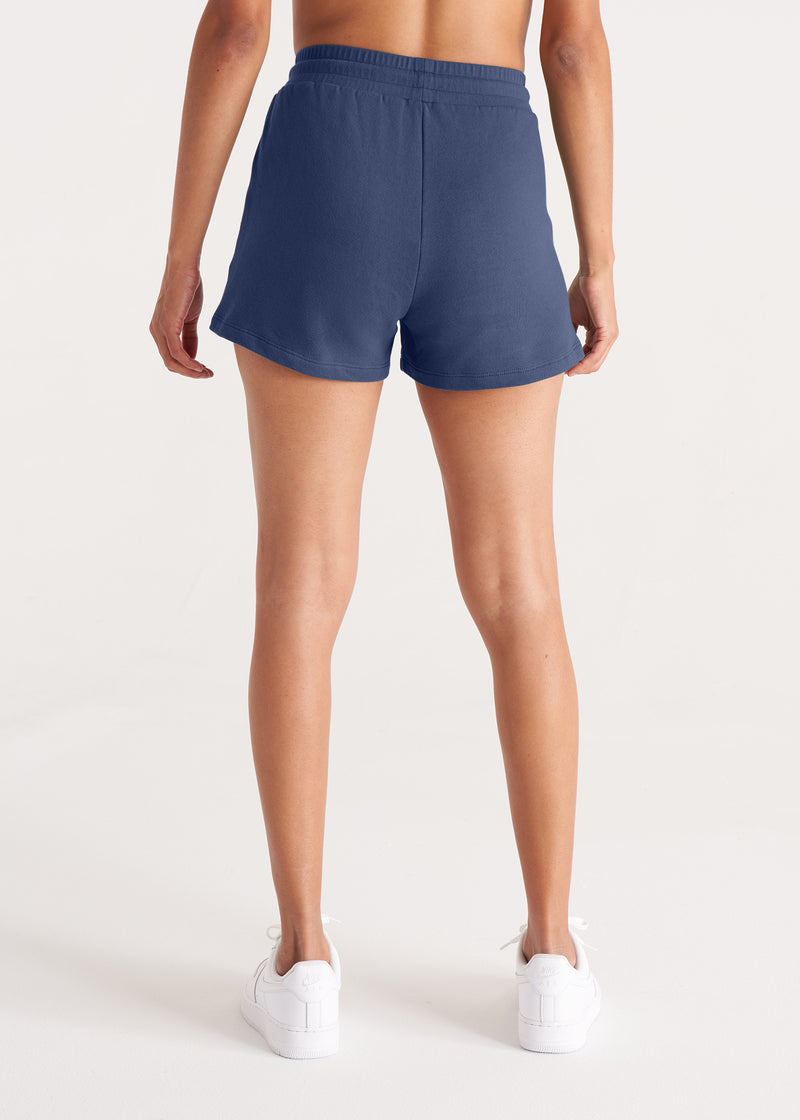 Woman in relaxed fit jogger shorts made of organic and recycled cotton French Terry with elastic waist, side pockets, and brass capped drawcords in color Midnight Navy, Back