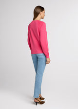 Woman in pink sweatshirt and blue denim with her hands at her side. Back.