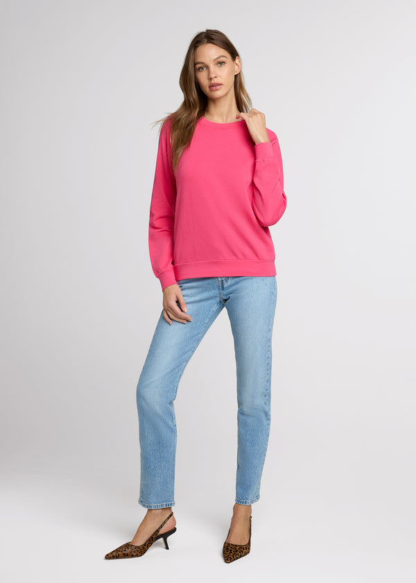 Woman in pink sweatshirt and light denim with one hand at her sweatshirt neckline and the other at her side wearing leopard print heels. Front. 