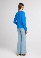Woman in blue raglan sweatshirt and light denim jeans with her hands at her side. Back.