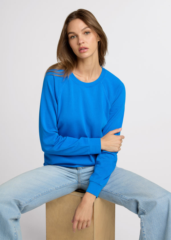 Woman in blue raglan sweatshirt and light denim jeans sitting on a wooden block as she crosses one hand across her body and the other at her side. Front. 