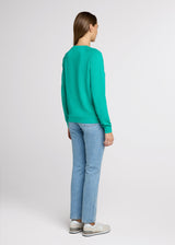 Woman in green sweatshirt and blue jeans standing with her hands at her side. Back.