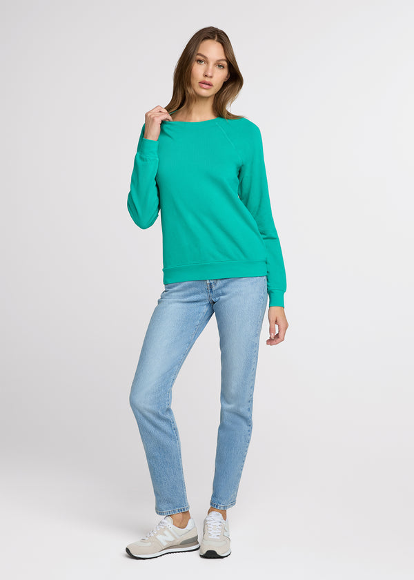Woman in green sweatshirt and blue jeans standing with one hand pulling at the collar of her sweatshirt and the other hand at her side. Front. 