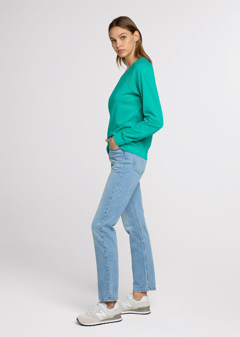 Woman in green sweatshirt and blue denim jeans standing to the side with one hand in her pant pocket and the other at her side. Side. 