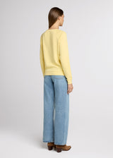 Woman in yellow sweatshirt and blue denim standing with her hands at her side. Back.
