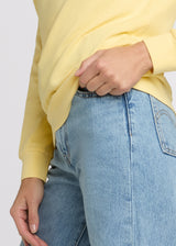 Woman in yellow sweatshirt and blue denim with one hand pulling up the hem of her sweatshirt while the other is at her side. Detail.