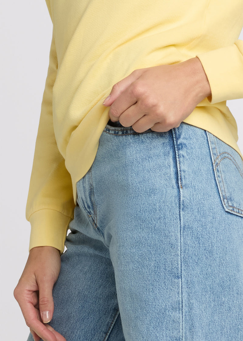 Woman in yellow sweatshirt and blue denim with one hand pulling up the hem of her sweatshirt while the other is at her side. Detail.