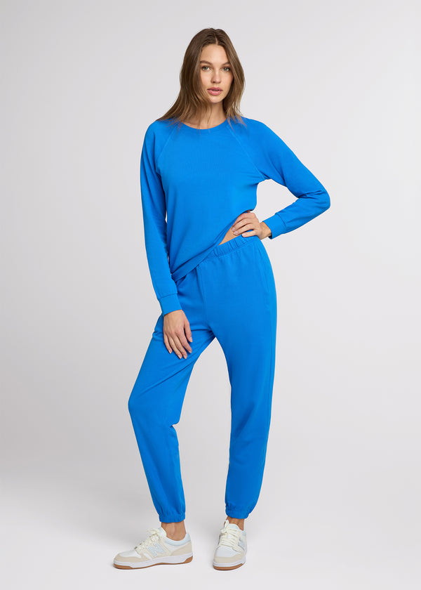 Woman in blue sweatpant and blue sweatshirt with her hand on her hip and the other at her side. 