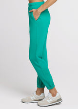 Woman in green sweatpant standing with her hands in her pocket. Side.