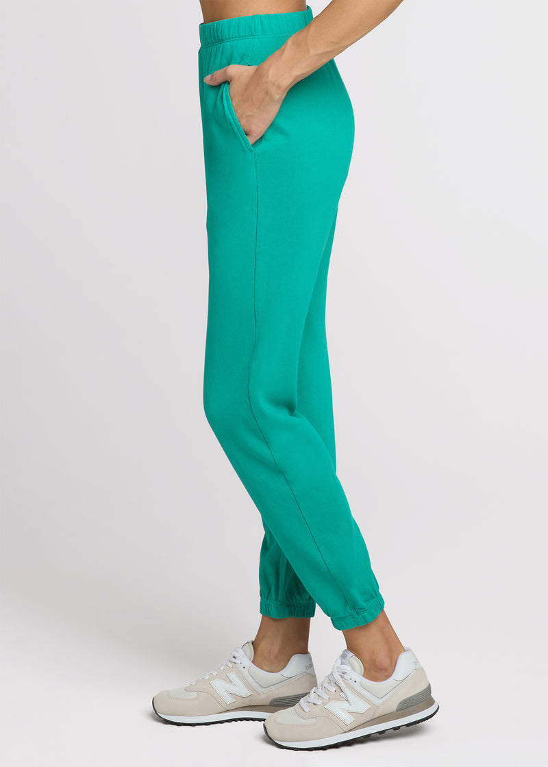 Woman in green sweatpant standing with her hands in her pocket. Side.