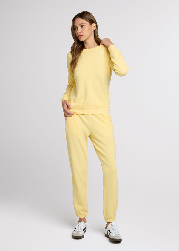 Woman in yellow sweatpant and yellow sweatshirt standing with one hand on her hip and the other pulling on the collar of her sweatshirt as she looks to the side. 