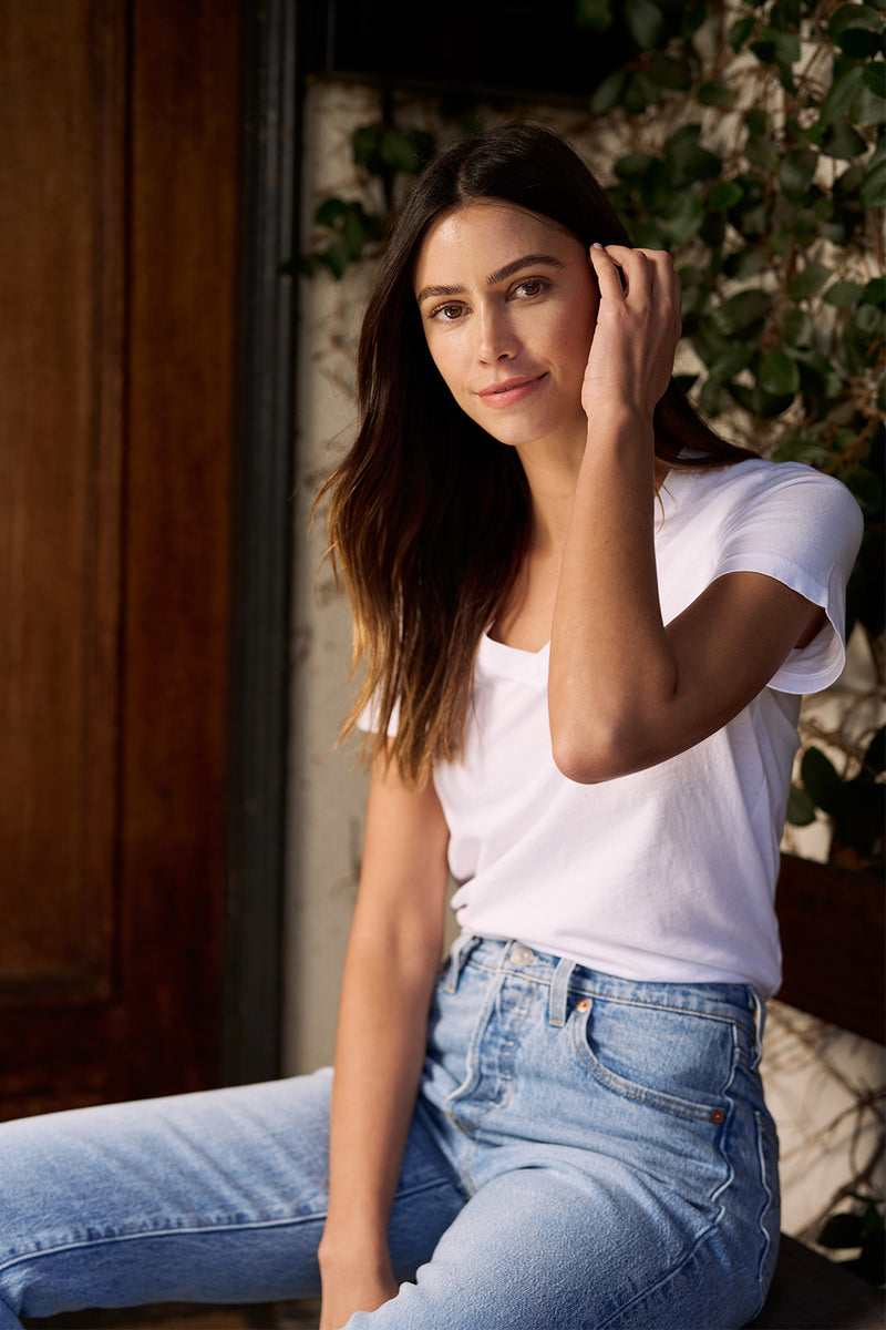 Women in white v neck tee and blue jeans sitting on a bench on the porch of a house with green ivy moving her hair behind her ears. 