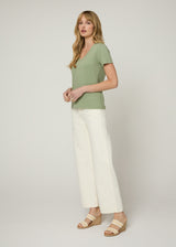 Woman in green v neck t-shirt and cream cropped denim jeans wearing white and nude wedges with her hands together at her front. Side.