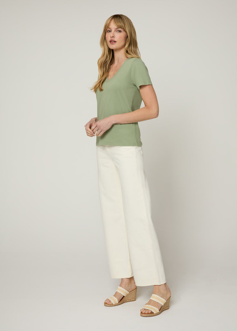 Woman in green v neck t-shirt and cream cropped denim jeans wearing white and nude wedges with her hands together at her front. Side.