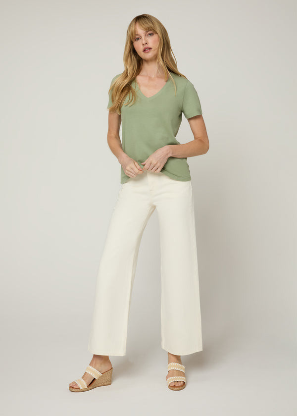 Woman in green v neck t-shirt with cream cropped denim pants and white and tan wedge heels holding the bottom of the t-shirt. Front. 