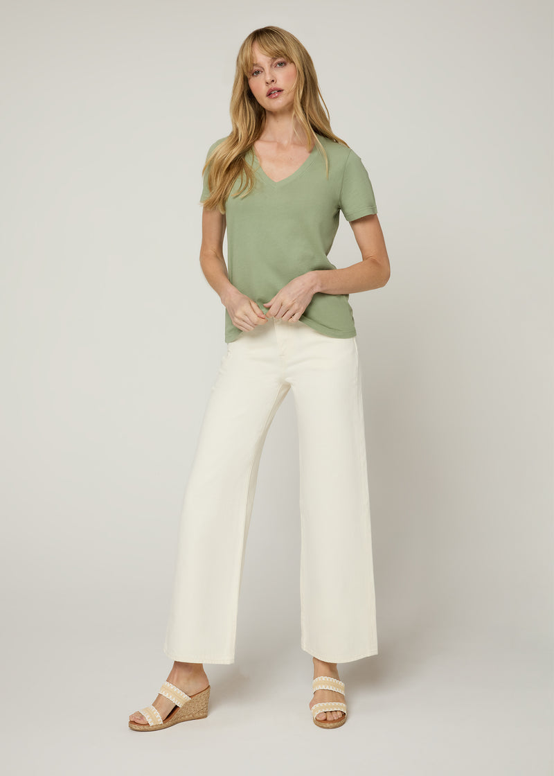 Woman in green v neck t-shirt with cream cropped denim pants and white and tan wedge heels holding the bottom of the t-shirt. Front. 