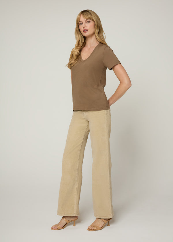 Woman in brown v neck t-shirt and tan jeans wearing nude heels with her hands behind her back. Side. 
