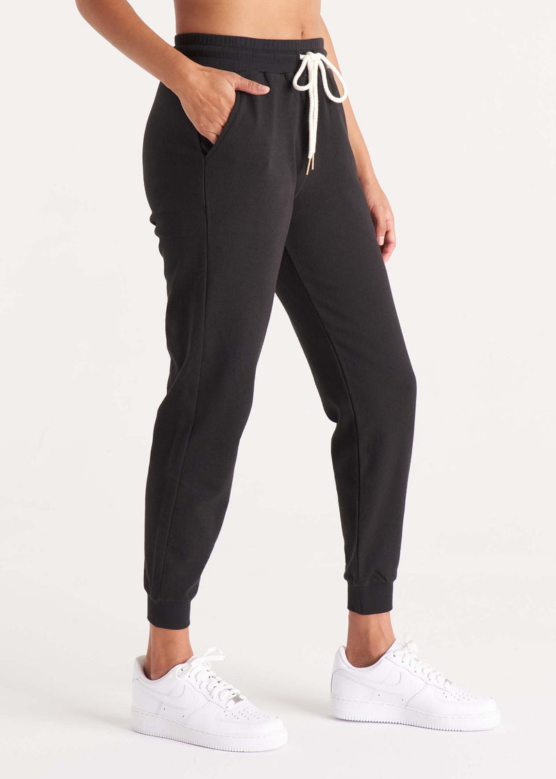 Woman in relaxed fit, mid-rise jogger pant with an elastic waist, pockets at sides, and brass capped drawcords in organic and recycled cotton French Terry in color onyx black, side image.
