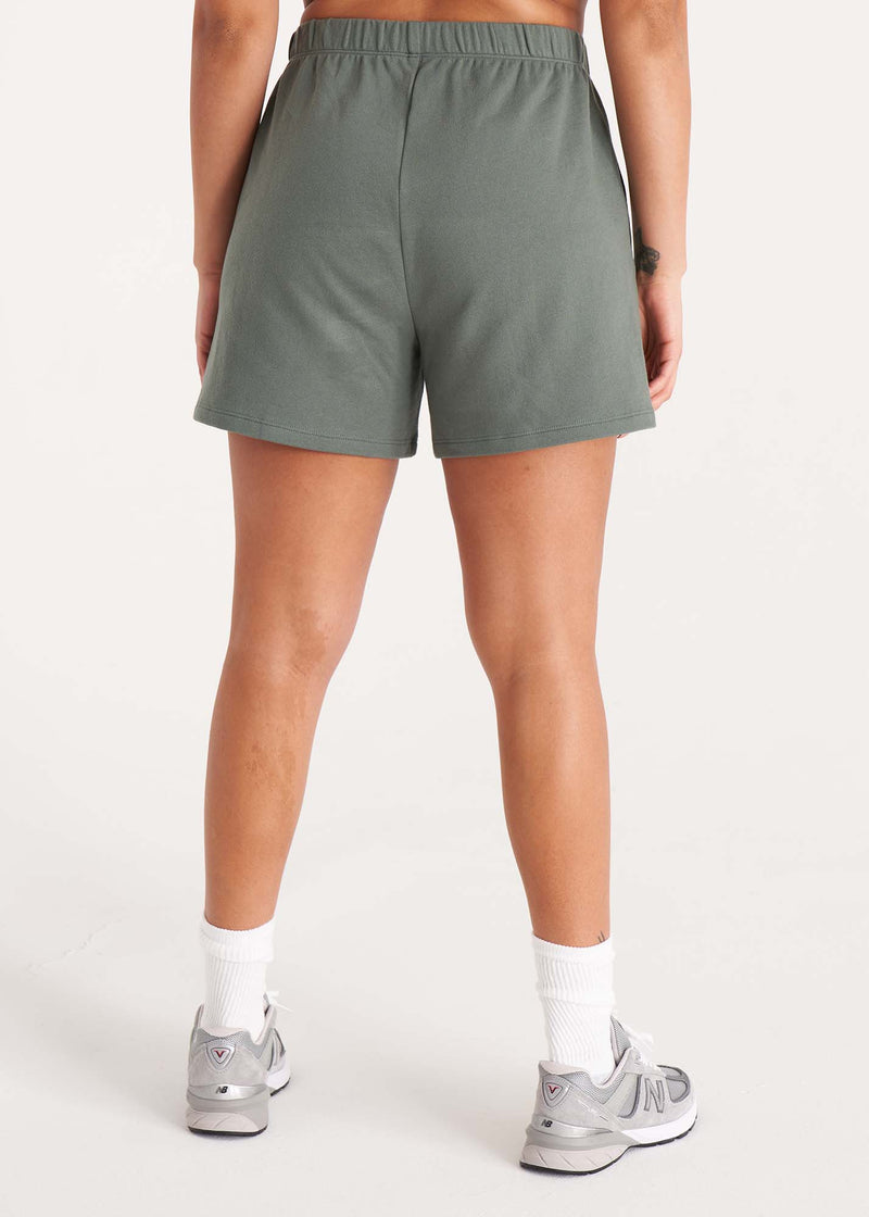 Woman in relaxed fit sweat shorts with elastic waist, side pockets, and interior drawcords made of organic and recycled cotton French Terry in color Thyme Green, Back