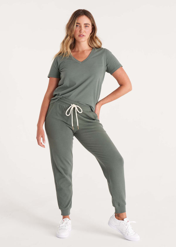 Woman in relaxed fit, mid-rise jogger pant with an elastic waist, pockets at sides, and brass capped drawcords in organic and recycled cotton French Terry in color thyme green paired with a v neck tee