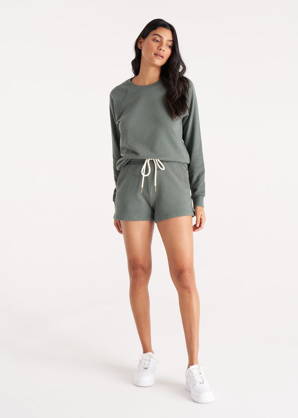 Woman in relaxed fit jogger shorts made of organic and recycled cotton French Terry with elastic waist, side pockets, and brass capped drawcords in color Thyme Green, Front 