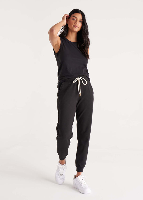 Woman in relaxed fit, mid-rise jogger pant with an elastic waist, pockets at sides, and brass capped drawcords in organic and recycled cotton French Terry in color onyx black paired with a tank top